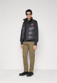 Black puffer vest with high collar and red logo, worn over a black sweatshirt, paired with tan pants and black slip-on shoes. Black beanie and sunglasses.