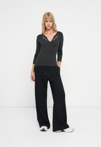 Top grigio in maglia con scollatura a V profonda e ricami a forma di ali; abbinato a pantaloni neri a gamba larga e sneakers bianche. Texture complessiva liscia.