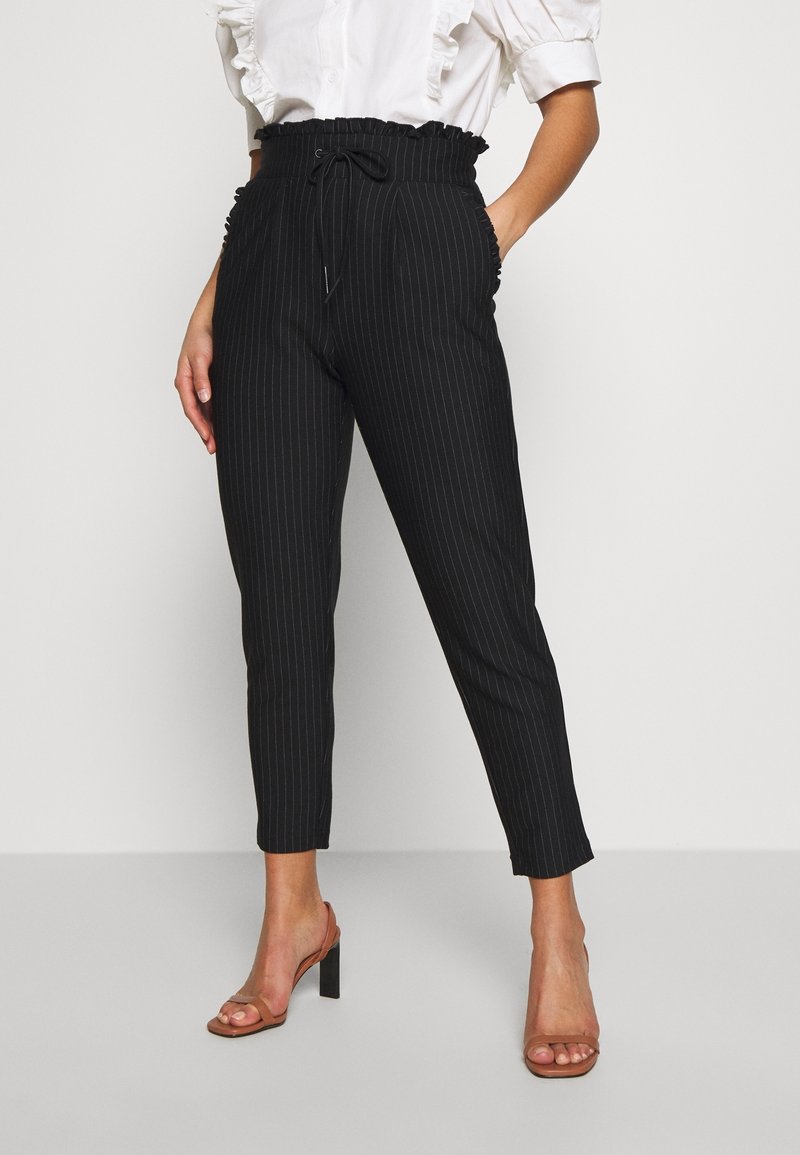 Femme portant un pantalon papier à rayures noires taille haute avec ceinture nouée, associé à un chemisier blanc à volants et des sandales marron à talons.