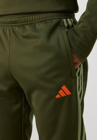 Olivza zaļas sporta bikses ar elastīgu jostasvietu, sānu kabatām un trim vertikālām gaiši zaļām svītrām. Raksturīgs sarkans Adidas logo.