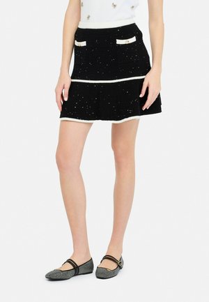 Jupe évasée noire avec des bordures blanches, dotée de deux poches avant ornées de boutons dorés et d'un tissu texturé avec des embellissements dispersés.