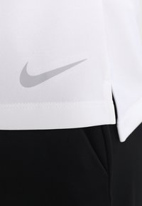 Bílá košile Nike s texturovaným povrchem, na boku s šedým logem. Černé sportovní kalhoty jsou částečně viditelné pod košilí.