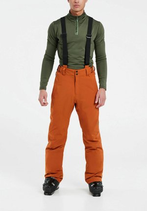 Pantalons de ski avec bretelles noires réglables, en tissu orange, fermeture à bouton et jambes larges. Portés avec des bottes de ski noires.