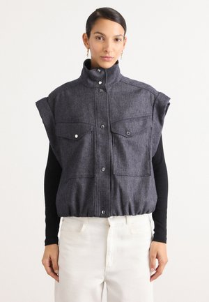Graue Wollweste mit hohem Kragen, Druckknöpfen und zwei Brusttaschen. Kurze Ärmel und geraffter Saum. Getragen über einem schwarzen Langarmoberteil.