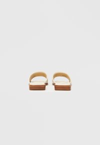 Dos sandalias vistas desde atrás con parte superior color crema y tacones gruesos de madera. Las tiras son redondeadas y presentan una textura acolchada.