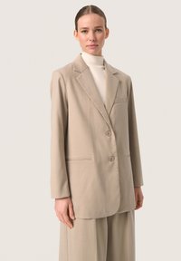 Beige blézer kerekre hajtott gallérral, két gombbal és egy egynyílású mellzsebbel; sima anyagból készült; magas nyakú toppal párosítva.