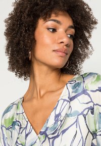 Vestido floral com decote em V, feito de um tecido leve. As cores incluem verde e azul sobre um fundo branco com padrões em linha.