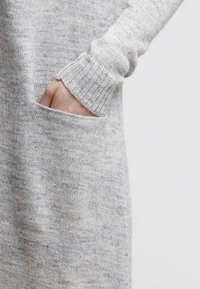 Ljust grå stickad tröja med ribbade ärmslut, med en framficka på sidan. Mjuk textur, avslappnad passform, synliga sömnadsdetaljer.