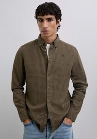 Camisa de cuadros marrón y verde con cuello puntiagudo, mangas largas y un bolsillo frontal; llevada sobre una camiseta blanca.