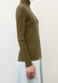 Personne portant un haut à manches longues côtelé olive avec une demi-fermeture éclair, associé à une jupe en denim bleu clair, vue de côté.