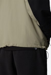 Beige og sort jakke med en glat, let tekstur; har en indsnøret bundkant og sorte fleecebukser under.