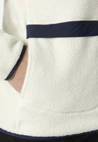 Suéter de forro polar crema con panel y ribete en azul marino, que presenta una textura suave y un bolsillo lateral. Detalles de costura visibles a lo largo de los bordes.