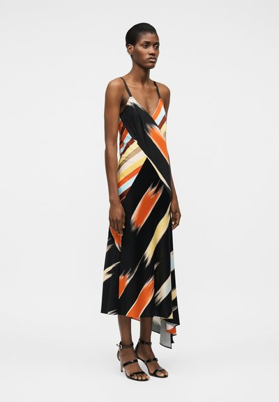 Γυναίκα που φοράει μίντι φόρεμα χωρίς μανίκια με πολύχρωμες ρίγες, ασύμμετρο τελείωμα και μαύρα σανδάλια με τακούνι, στέκεται μπροστά σε μονόχρωμο φόντο.