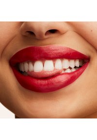 Jasnoczerwona szminka na pełnych ustach, podkreślająca gładką teksturę. Widać białe zęby, z bliskim ujęciem na okolicę ust.