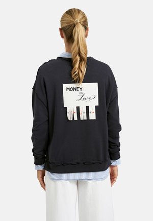 Person with blonde ponytail wearing black sweatshirt with a paper sign reading "Money or Love?" and tear-off tabs with "$" and hearts.