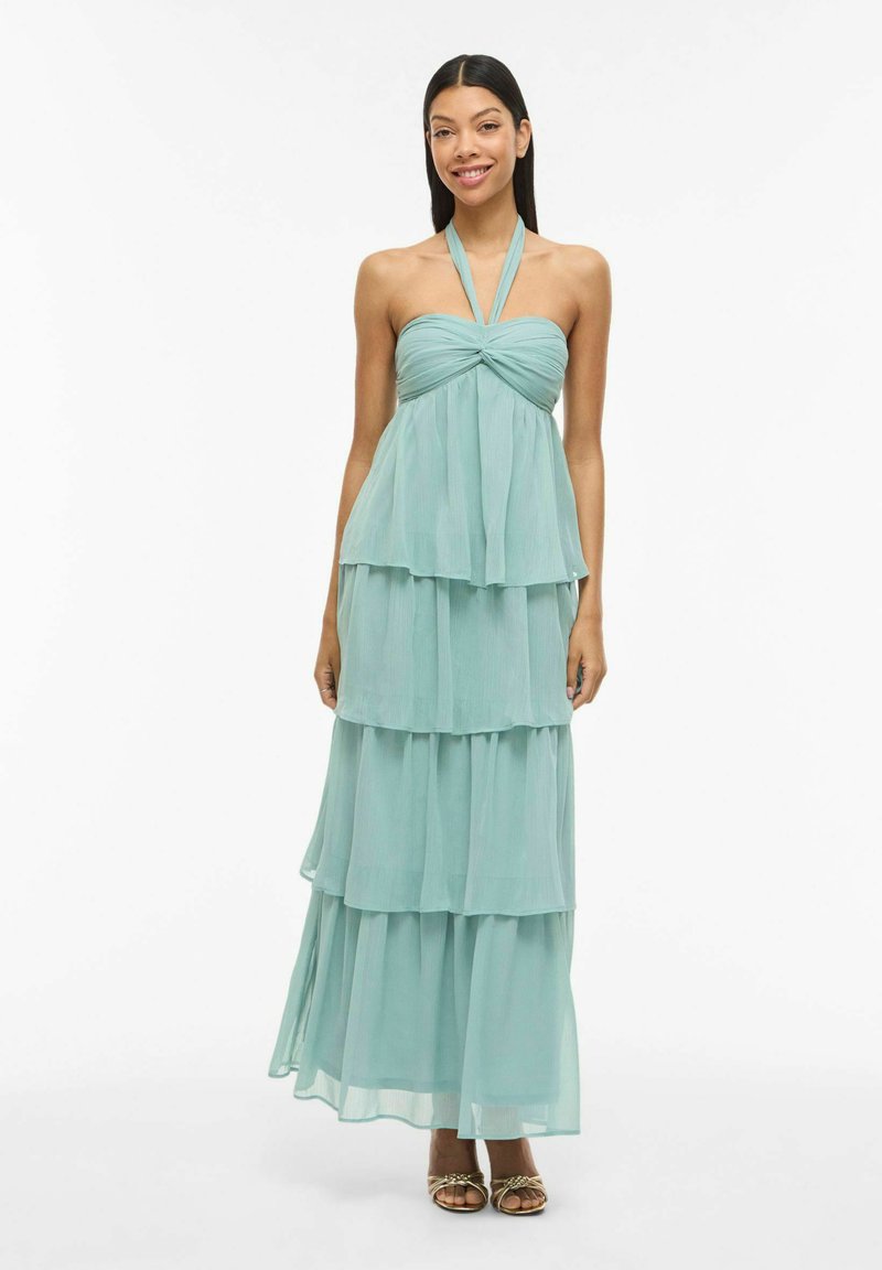 Femme debout portant une robe maxi turquoise claire, sans bretelles et à plusieurs niveaux, ainsi que des sandales dorées à bout ouvert, souriante, les mains détendues le long du corps.