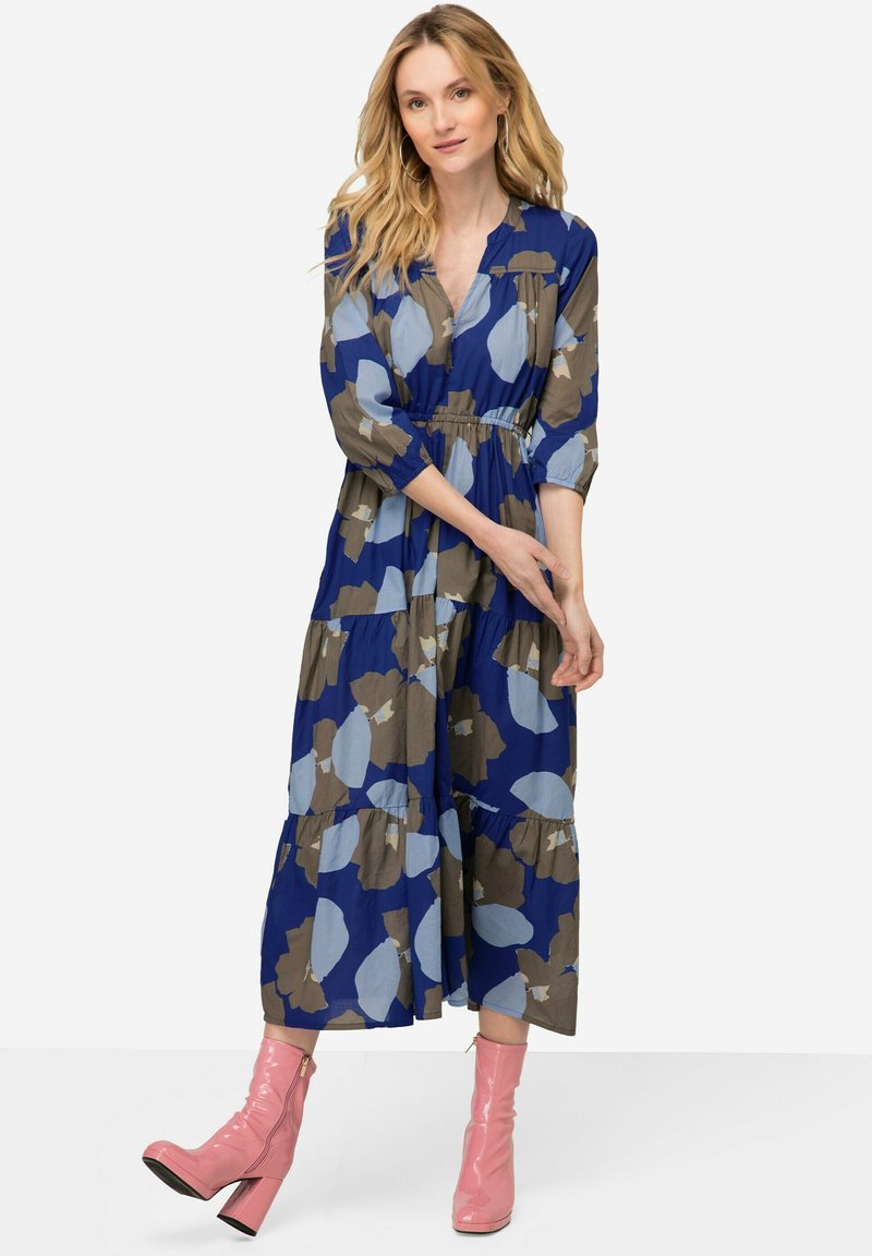 Vestido maxi floral con patrones en azul, azul claro y marrón, con escote en V, mangas tres cuartos y falda en capas. Combinado con botas de tobillo rosas.
