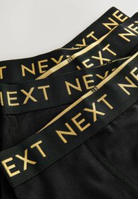 Crne gaćice s crnim pojasom na kojem se ističe zlatni "NEXT" logo, glatke teksture i uskog kroja. Uređene u urednom rasporedu.