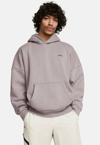 Felpa viola chiaro con cappuccio, dotata di tasca frontale e polsini a costine, realizzata in materiale morbido, indossata con pantaloni di colore chiaro.