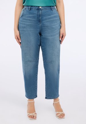 Vrouw die een blauwe denim mid-rise straight-leg jeans met knienaad draagt, gecombineerd met witte sandalen met hak en bandjes en een teal top.