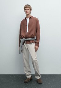 Chemise en lin marron, t-shirt blanc, pantalon beige, pull gris noué à la taille, chaussures marron, debout contre un fond clair.