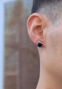 Boucles d'oreilles carrées noires en métal, avec quatre griffes en pointe. Vue sur l'oreille d'une personne contre un fond texturé.