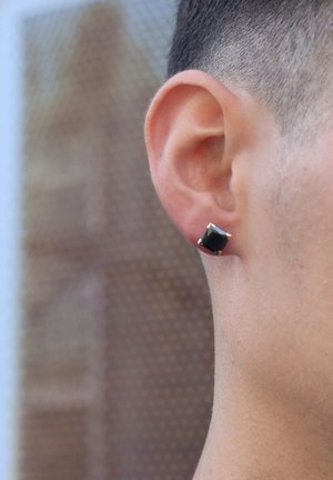 Schwarzes quadratisches Steck-Ohrstecker-Set aus Metall, mit vier spitzen Prong-Verzierungen. Auf dem Ohr einer Person vor einem strukturierten Hintergrund zu sehen.
