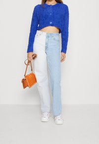 Modrý chlupatý krátký cardigan se zapínáním na knoflíky, spárovaný s džíny s rovnými nohavicemi ve světlém odstínu. Oranžová kabelka a bílé tenisky dokreslují outfit.