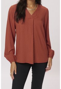 Rustfarvet blouse med et tekstureret mønster, V-udskæring og lange, elastiske ærmer. Båret sammen med tætsiddende sorte bukser.