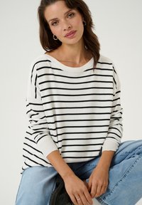 Pull rayé blanc et noir avec un large col et une coupe ample. Texture douce, poignets côtelés et manches relaxées associées à un jean bleu clair.