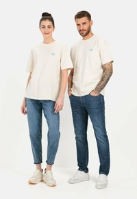 Cremefarbene Baumwoll-T-Shirts mit blauem Logo, kombiniert mit hellblauen Jeans und weißen Sneakers. Das T-Shirt hat eine lockere Passform und kurze Ärmel.