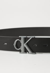 Cinto de couro preto com fivela prateada metálica apresentando as letras "ck." Inclui furos para ajuste de tamanho. Textura suave.