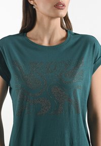 Žena v tyrkysovém tričku s abstraktními, třpytivými vzory. Její výraz je neutrální, s jemným make-upem a viditelnými náušnicemi.