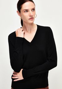 Zwarte V-hals trui, lange mouwen, geribde manchetten en zoom, zachte textuur, aansluitend design. Model poseert met één hand op haar hals.