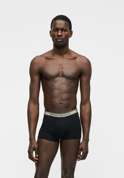 Μυώδης άνδρας στέκεται μπροστά, φορώντας μαύρα μπόξερ Emporio Armani με γκρι μέση, σε απλό γκρι φόντο.