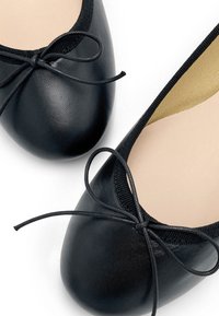 Ballerine nere realizzate in materiale sintetico liscio, con punta arrotondata e un delicato fiocco decorativo sulla tomaia. Soletta color beige chiaro visibile.