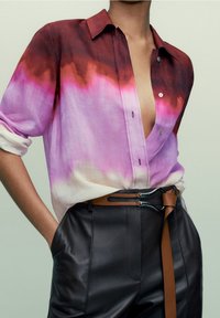 Camicia con gradiente colorato nelle tonalità di rosso, rosa e bianco; tessuto leggero, vestibilità rilassata, stile a bottoni, abbinata a pantaloni in pelle scura e una cintura marrone.