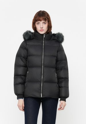 Vrouw in een zwarte pufferjack met een capuchon van bont, een rits aan de voorkant en ritszakken aan de zijkanten, gecombineerd met donkerblauwe jeans.