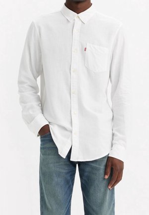 Mand iført en hvid langærmet skjorte med brystlomme og blå jeans, med den ene hånd i lommen, mod en ensfarvet hvid baggrund.