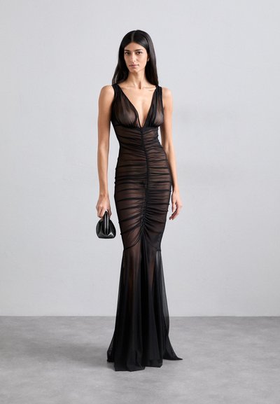 Schwarzes, durchsichtiges Abendkleid mit tiefem V-Ausschnitt und gerafftem Detail in der Mitte. Ausgestellt am Saum und mit dünnen Trägern. Hält eine kleine, strukturierte schwarze Tasche.