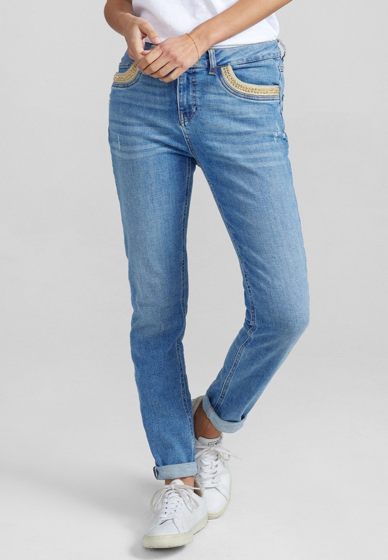 Femme portant un jean bleu clair avec des embellissements en perles dorées sur les poches avant, des baskets blanches et un haut blanc, debout les jambes croisées.