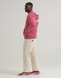 Felpa rosa con cappuccio e tasche frontali, abbinata a pantaloni beige a gamba larga. Calzature sono sneakers rosse con dettagli bianchi.