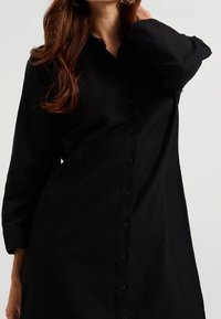 Svart skjortklänning i en mjuk tygkvalitet, med krage, långa ärmar och en avslappnad passform med knapprader längs framsidan.