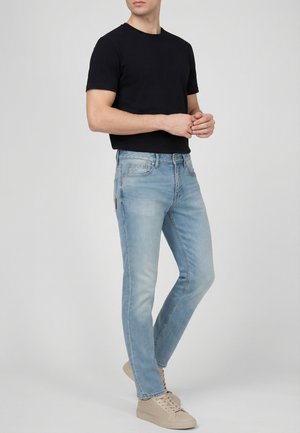 Homme portant un t-shirt noir uni à manches courtes, un jean slim bleu clair et des baskets beige, debout les mains jointes.