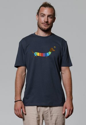Mann mit Bart und zurückgebundenem Haar trägt ein marineblaues T-Shirt mit einem bunten gestreiften Dackel-Motiv, beige Hose und ein schwarzes Armband und steht vor einem grauen Hintergrund.