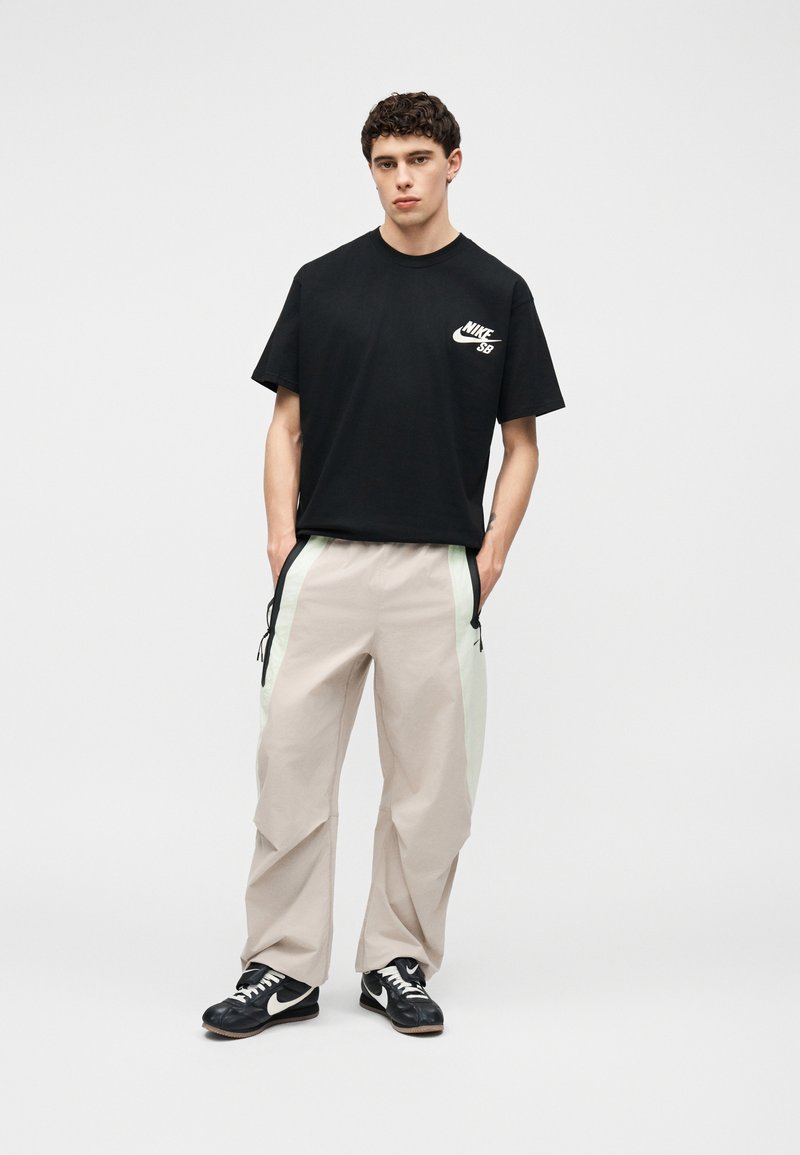 Jonge man draagt een zwart Nike SB T-shirt, losse beige trainingsbroek met lichte zijstrepen en zwarte Nike sneakers, staande met handen in de zakken.