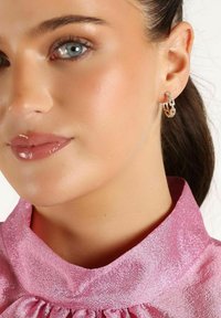 Guldörhängen med en sammanlänkad design, prydda med klara ädelstenar. Modell bär en rosa strukturerad blus med hög krage.