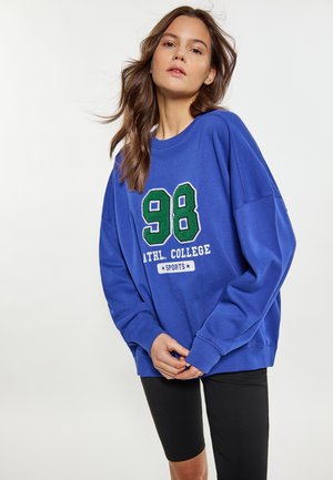 Vrouw die een blauwe trui draagt met de tekst "98 ATHL. COLLEGE SPORTS" en zwarte knielange legging, staand tegen een effen witte achtergrond.