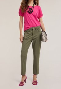 Fuksjowa bluzka z bufiastymi rękawami, oliwkowe dopasowane spodnie, różowe sandały na obcasie z paskami oraz torebka w szarym wężowym printie o fakturze. Odważne naszyjniki jako akcenty.