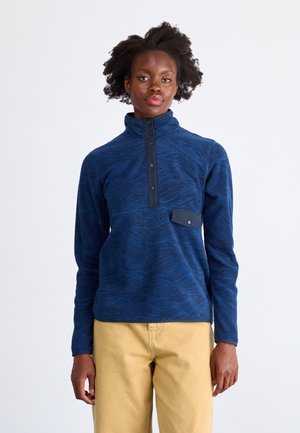 Polarowy sweter w granatowym kolorze z wzorem fal, z pół-zatrzaskiem, wysokim kołnierzem, kieszenią na piersi oraz ściągaczami na rękawach, zestawiony z jasnobeżowymi spodniami.